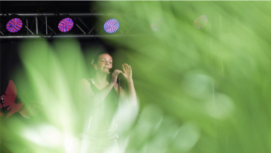 Dame på scenen som synger, med grønne palmeblader i forgrunn av bildet